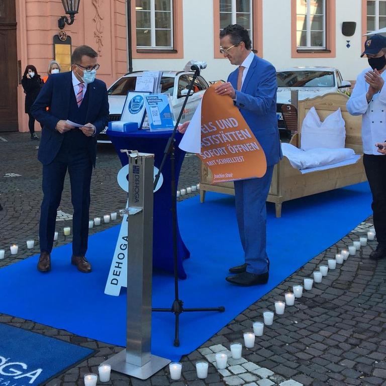 Vor simulierter Hotel-Kulisse mit Bett und Teppich: Joachim Streit, Spitzenkandidat der Freien Wähler in Rheinland-Pfalz, entrollt ein Plakat mit seiner Wahlkampf-Forderung: „Hotels und Gaststätten sofort öffnen – mit Schnelltest“.