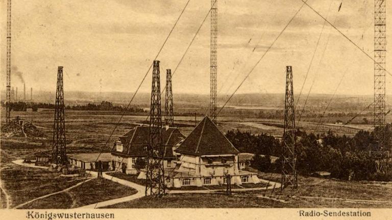Eine historische Postkarte zeigt einige Gebäude, zwischen denen hohe Masten stehen, die über lang gespannte Seile gesichert sind.