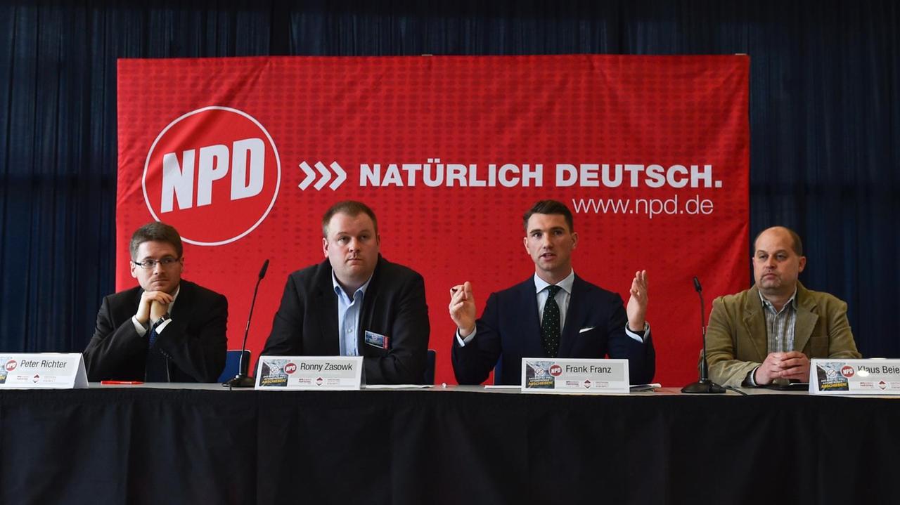 Man sieht von links nach rechts die NPD-Parteifunktionäre Peter Richter, Ronny Zasowk, den Parteivorsitzenden Frank Franz und Klaus Beier an einem Tisch vor einer roten Fahne.