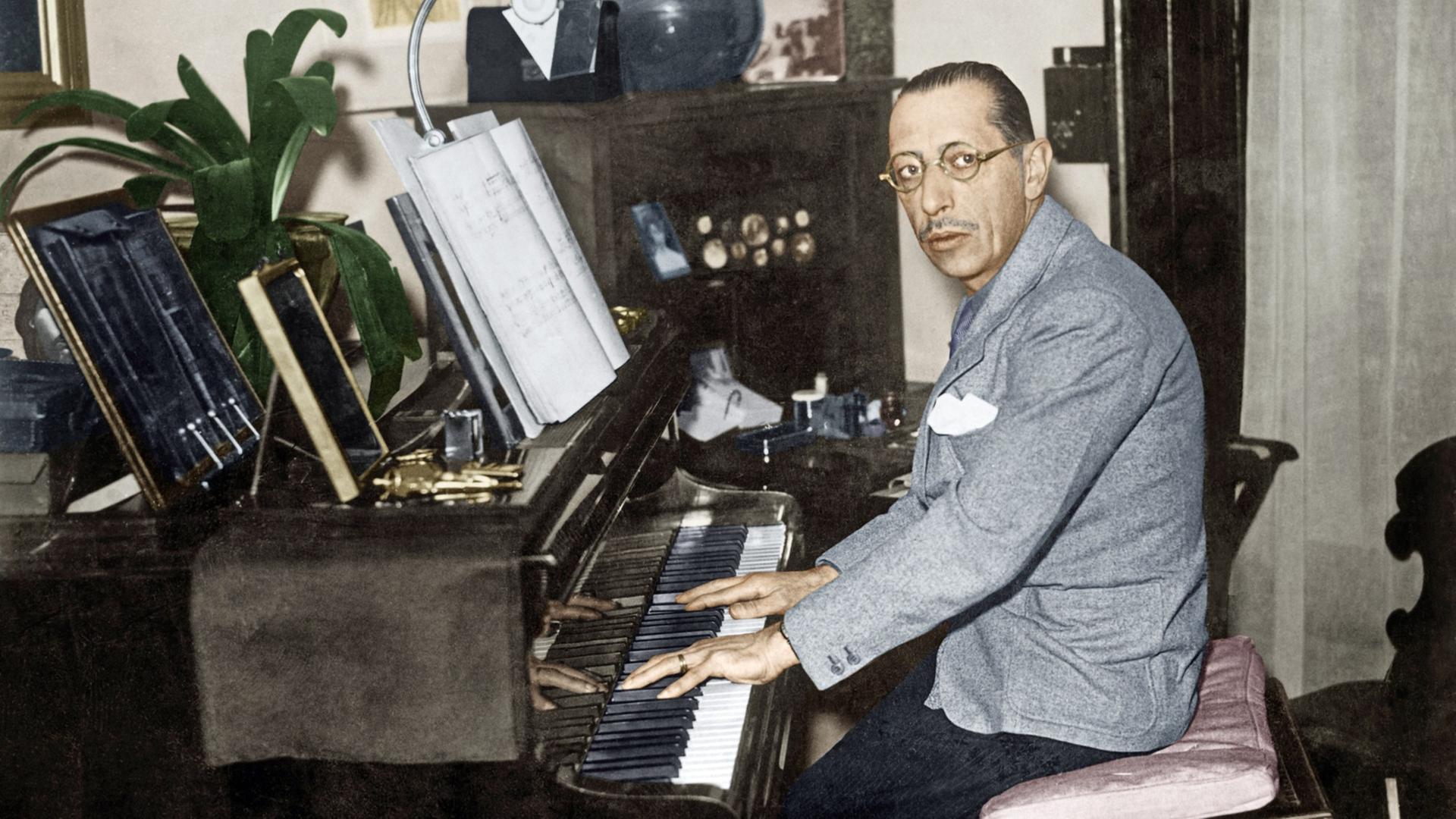Ein Mann mit streng zurückgekämmten Haaren sitzt am Klavier und blickt durch eine runde Brille nach links in die Kamera. Er trägt hellgraues Jackett mit weißem Einstecktuch und schwarze Cordhose.