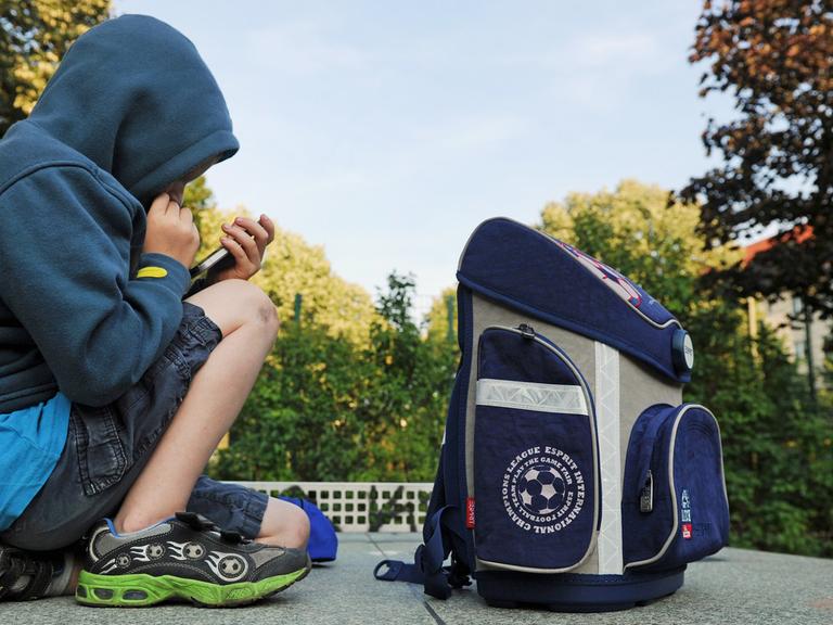 Ein Junge sitzt mit seinem Schulranzen auf einer Tischtennisplatte und spielt auf einem Smartphone.