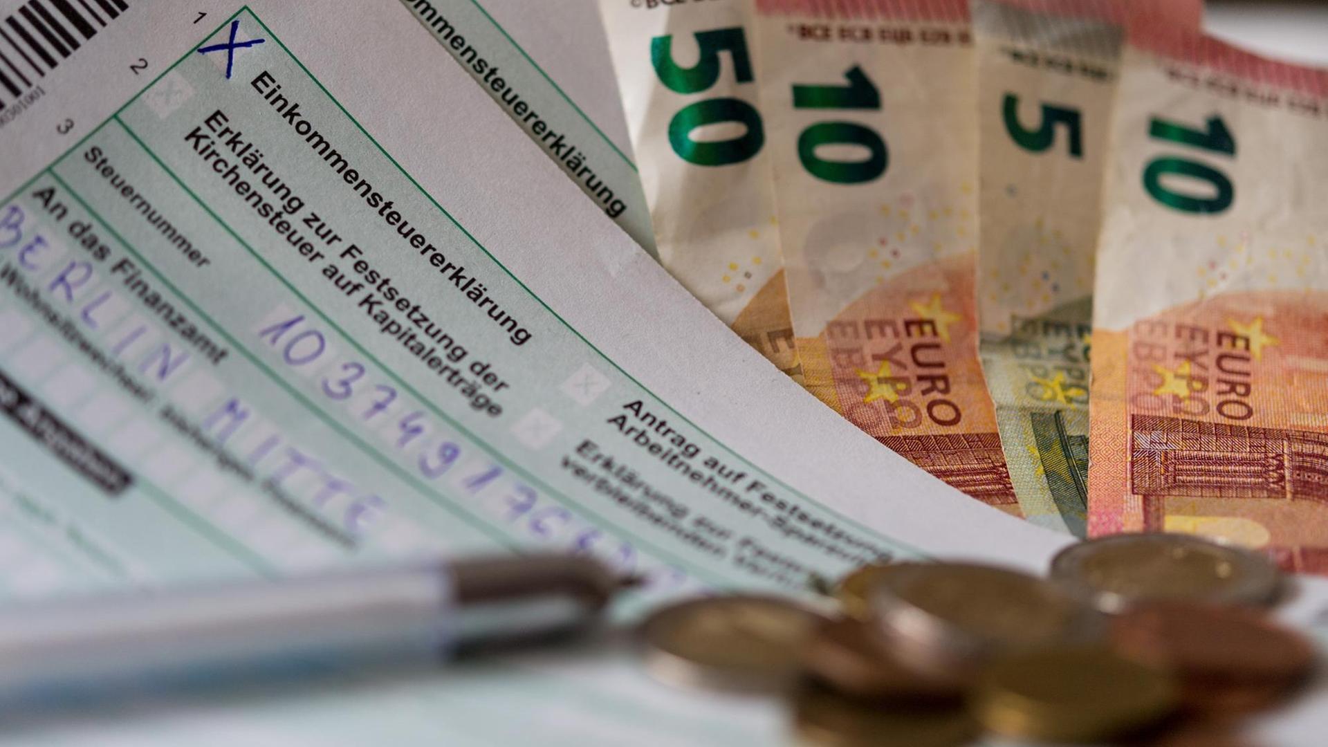 Ein Formular für eine Steuer-Erklärung liegt mit Geld-Scheinen und Münzen auf einem Tisch.