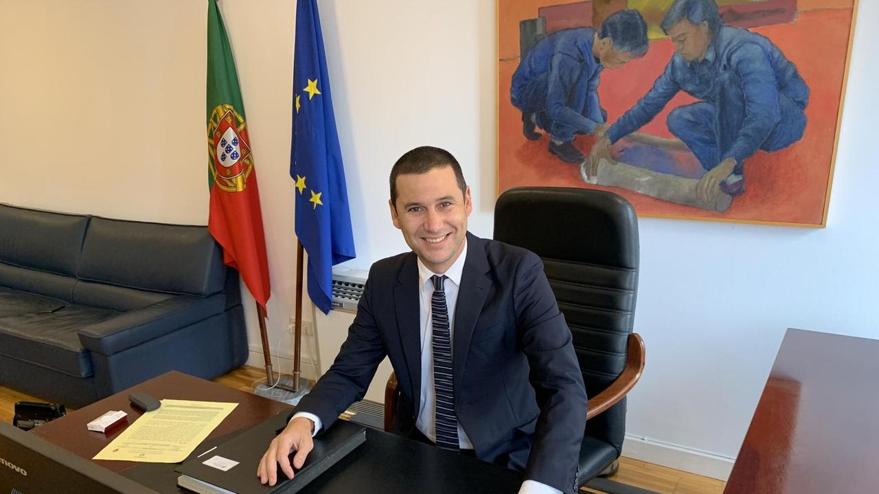 Ein junger Mann sitzt in die Kamera strahlend in seinem Büro am Schreibtisch, hinter ihm die Fahne Europas und Portugals.