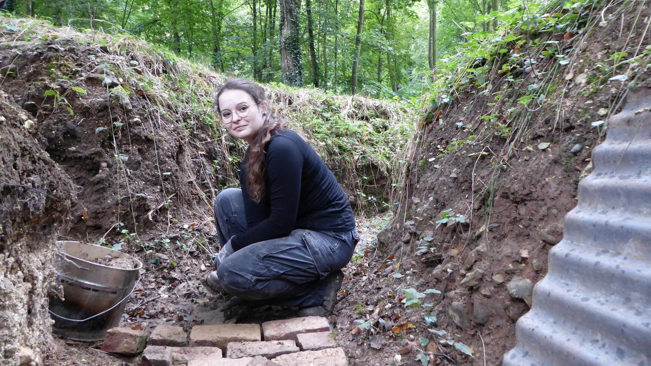 Eine Frau mit Arbeitskleidung sitzt in einem Graben und lächelt in die Kamera.