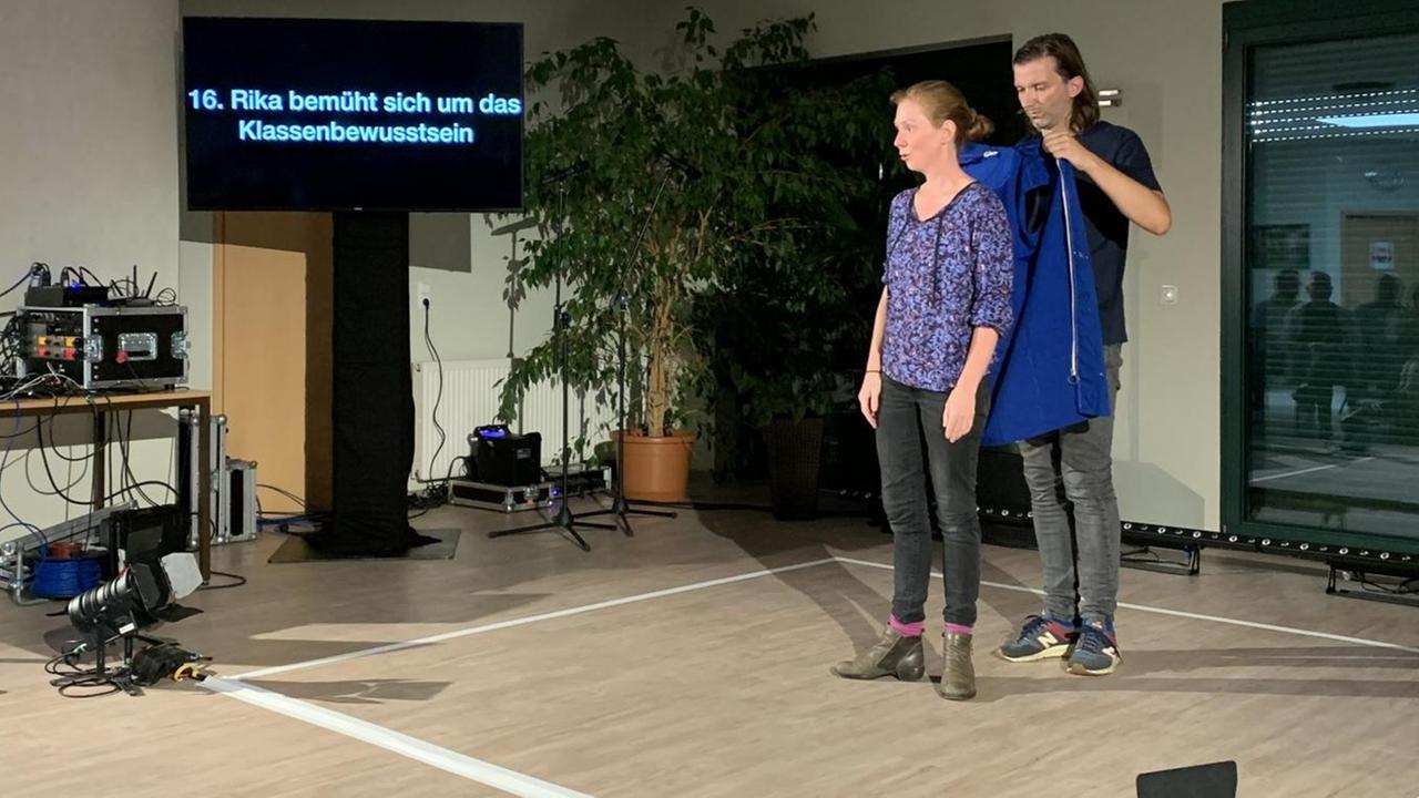 Rika Weniger steht in einem Raum. Auf dem Boden ist mit Klebestreifen ein großes Quadrat abgeklebt. Darin steht sie und ihr Bühnenpartner hilft ihr in eine blaue Jacke.