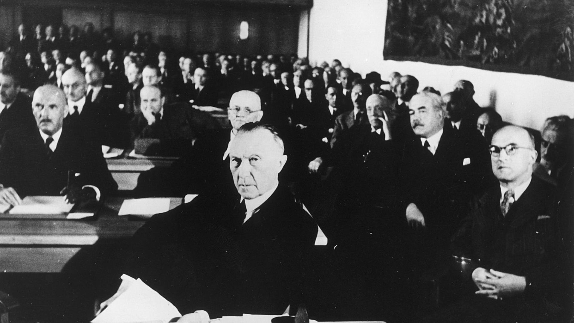 Das schwarz-weiße Bild zeigt die Mitglieder an Tischen sitzend. Im Vordergrund Adenauer. Das schwarz-weiße Bild zeigt die Mitglieder an Tischen sitzend. Im Vordergrund Adenauer.