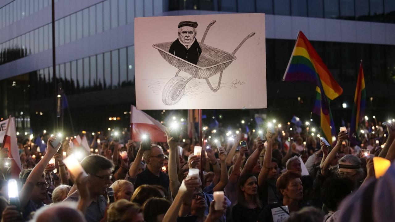 Hunderte Protestierende halten während der Dämmerung brennende Kerzen in den Händen. Auf einem Plakat ist eine Zeichnung von Jarosław Kaczyński in einer Sackkarre zu sehen.