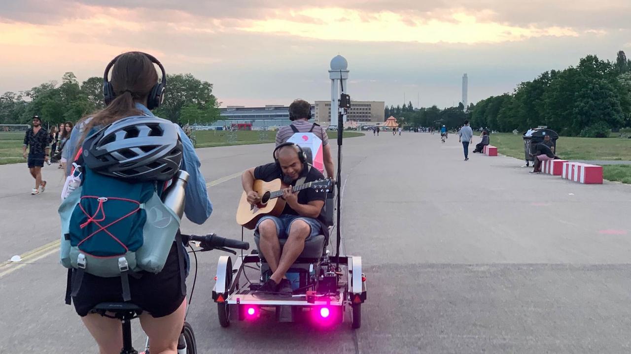 Eine Fahrradfahrerin mit Kopfhörern auf dem Tempelhofer Feld, vor ihr fährt ein Radfahrer mit Anhänger, auf dem ein Musiker Gitarre spielt.