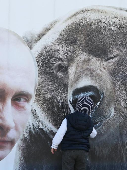 Ein kleiner Junge inspiziert eine Plakatwand mit einem zwinkernden russischen Präsidenten Wladimir Putin und einem Bären, sowie einem T-Shirt mit der Aufschrift: "Ich bin ein Freund von Putin".