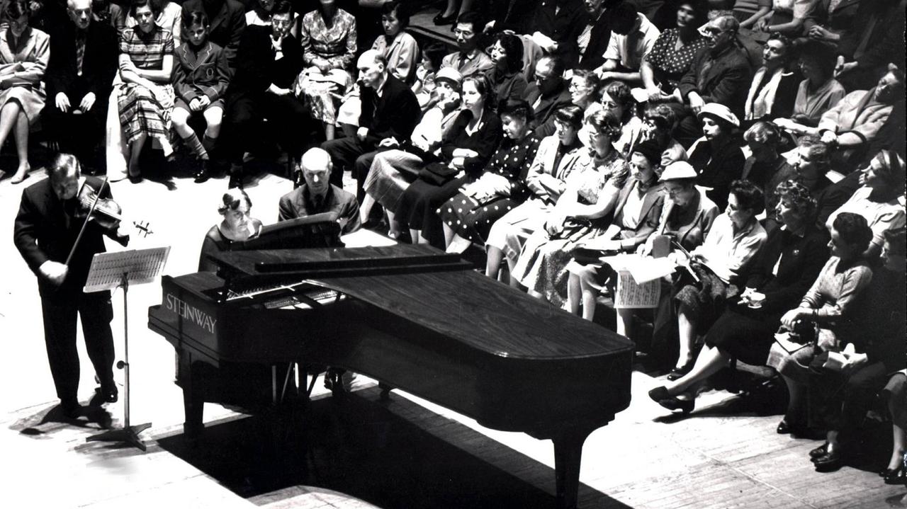 Schwarz-Weiß-Bild, links steht der Geiger Isaac Stern vor einem Notenpult, rechts neben ihm, an einem großen schwarzen Steinwayflügel, sitzt die Pianistin Myra Hess. Das Publikum sitzt drumherum ebenerdig und ganz nah