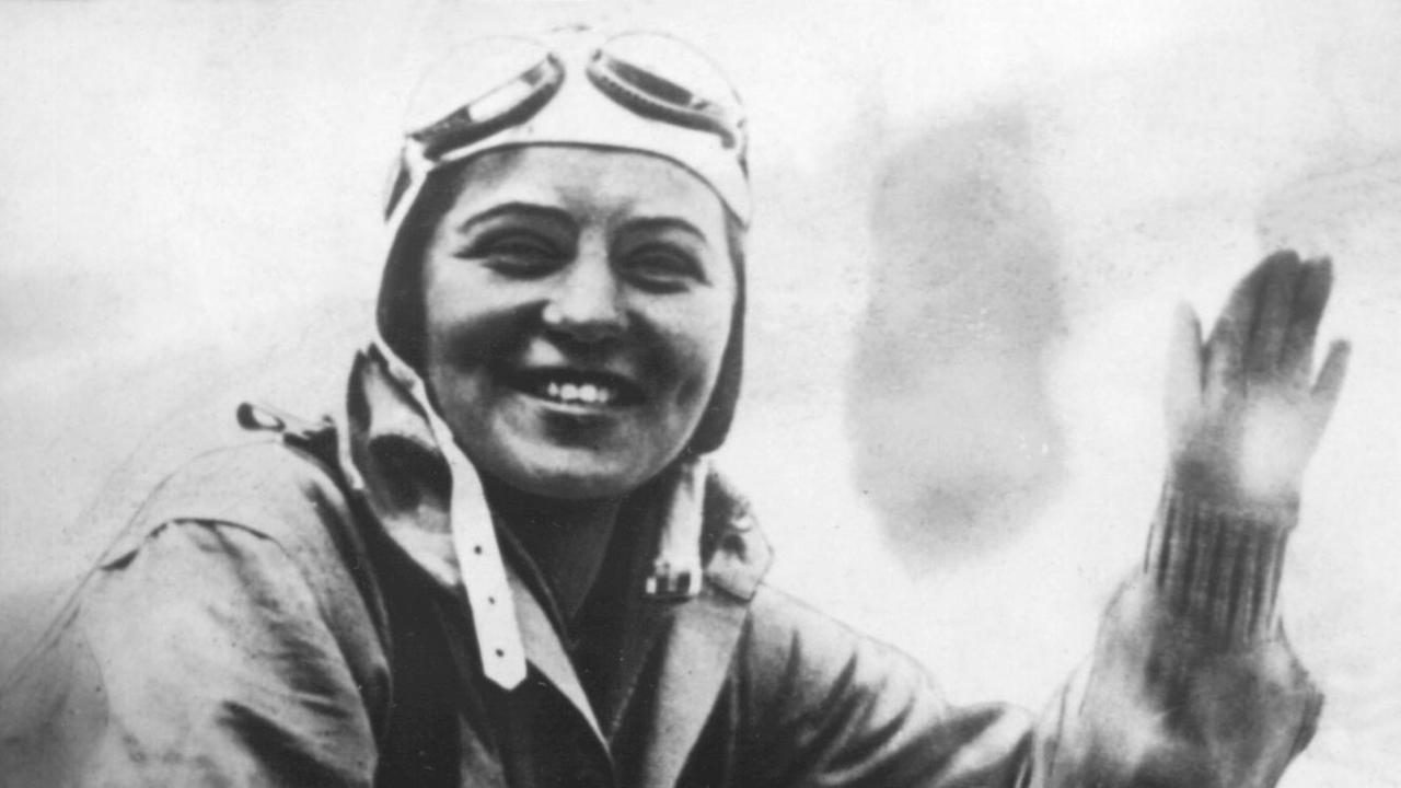 Ein Schwarz-Weiß-Foto sus den 1930er-Jahren zeigtdie Fliegerin Elly Beinhorn in die Kamera lachend und winkend im Cockpit eines Kleinflugzeugs