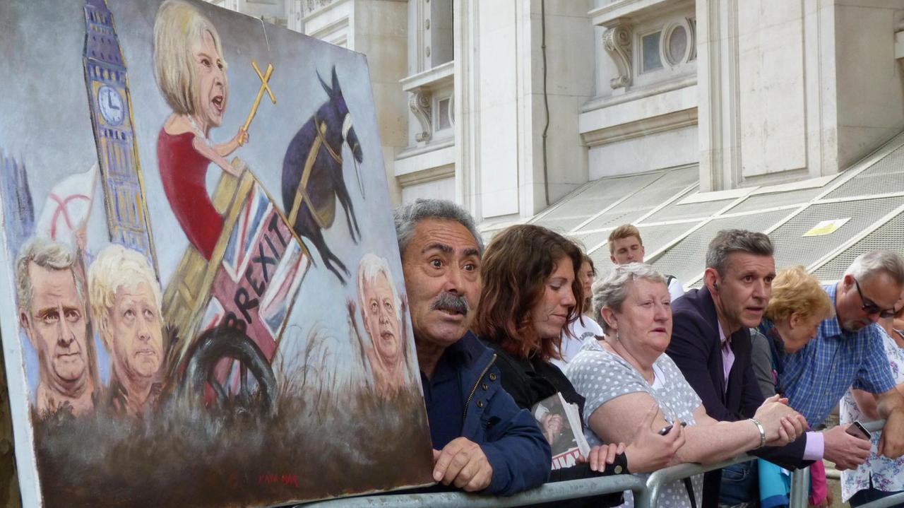 Menschen warten vor der Downing Street 10 auf Premierministerin Theresa May. Ein Mann hält eine Karrikatur von May in der Hand,  die das Thema Brexit thematisiert.