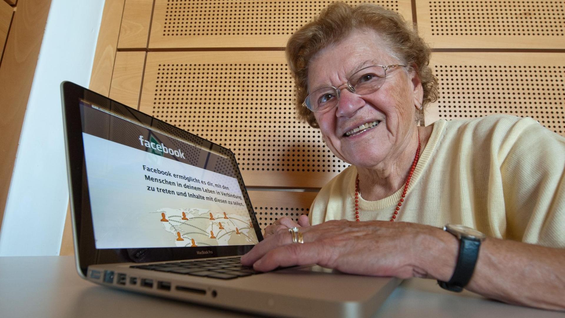 Seniorin sitzt vor einem Laptop mit der Startseite von Facebook auf dem Monitor.