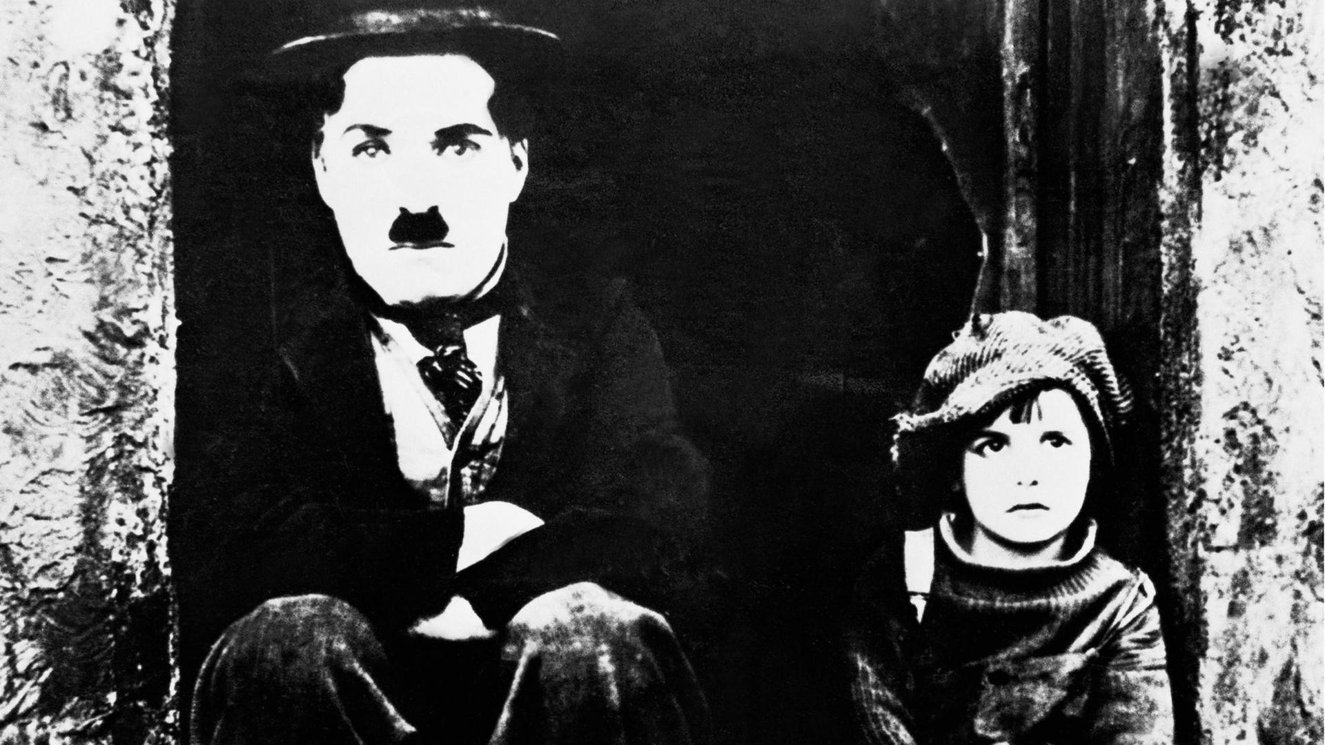 Ein schwarz-weißes Film-Still zeigt: Charlie Chaplin (links) - mit seiner typischen Melone und Schnauzer - als Tramp und Jackie Coogan als Kid in "THE KID" von 1921. Beide sitzen auf einem Treppenabsatz und blicken frontal al in die Kamera.