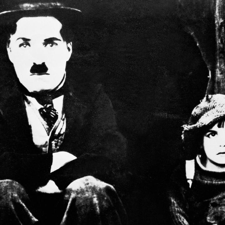 
Ein schwarz-weißes Film-Still zeigt:
Charlie Chaplin (links) - mit seiner typischen Melone und Schnauzer - als Tramp und Jackie Coogan als Kid in "THE KID" von 1921. Beide sitzen auf einem Treppenabsatz und blicken frontal al in die Kamera.