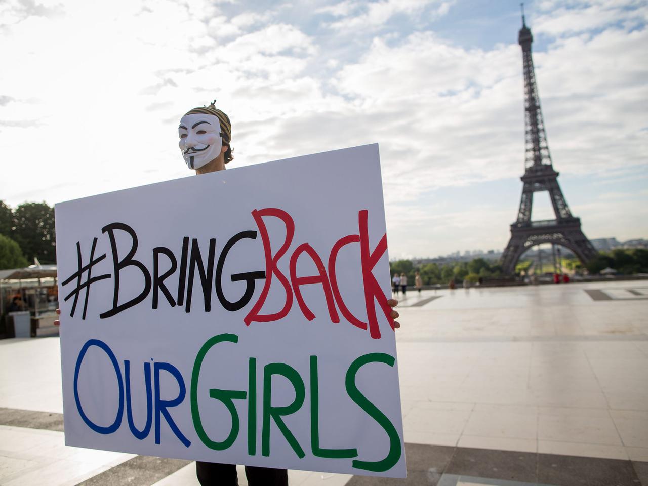 Ein maskierter Mann hält ein Schild mit der Aufschrift "Bring back our girls", in dem Hintergrund der Eiffelturm.