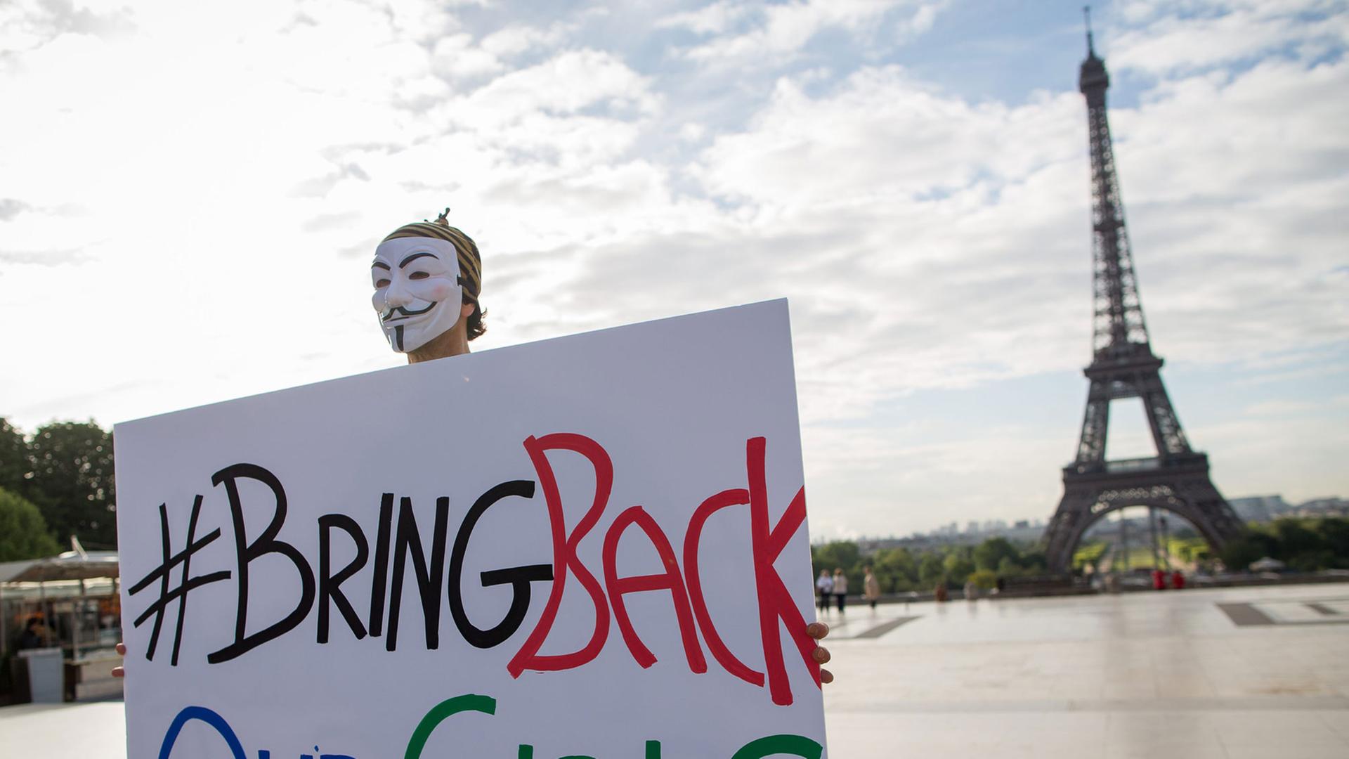 Ein maskierter Mann hält ein Schild mit der Aufschrift "Bring back our girls", in dem Hintergrund der Eiffelturm.