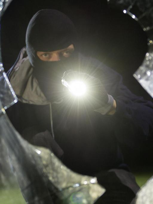 Das Bild zeigt einen fiktiven Einbrecher, er schaut mit Taschenlampe durch eine geborstene Scheibe.