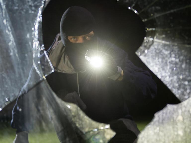 Das Bild zeigt einen fiktiven Einbrecher, er schaut mit Taschenlampe durch eine geborstene Scheibe.