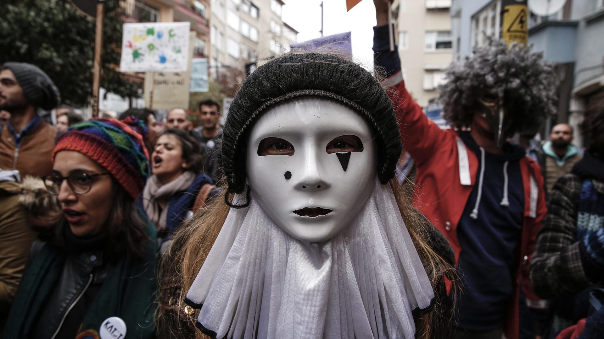 Menschen gehen demonstrierend durch eine Straße. In der Mitte des Bildes ist eine Frau oder ein Mann mit einer weißen Maske auf dem Gesicht.