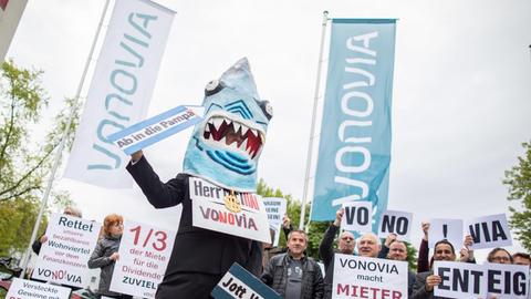 16.05.2019, Nordrhein-Westfalen, Bochum: Ein Demonstrant in einem Hai-Kostüm mit einem Schild um den Hals "Herr Miethai Vonovia" steht vor der Vonovia Hauptversammlung vor dem Ruhrcongress. Weitere Demonstranten stehen dahinter. Foto: Marcel Kusch/dpa | Verwendung weltweit