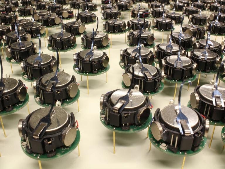Hunderte von kleinen, runden Robotern auf jeweils drei dünnen Beinen stehen auf einer Fläche herum.