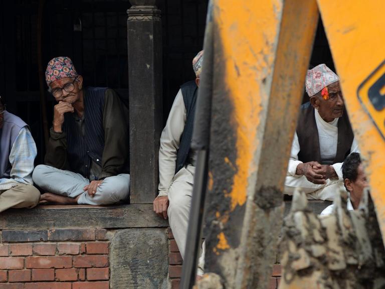 Nepalesen sitzen auf einer Mauer vor der ein Bagger steht.