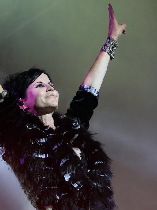 Dolores O’Riordan steht mit nach oben gereckten Armen auf der Bühne, in einer Hand hält sie ein Mikrofon.