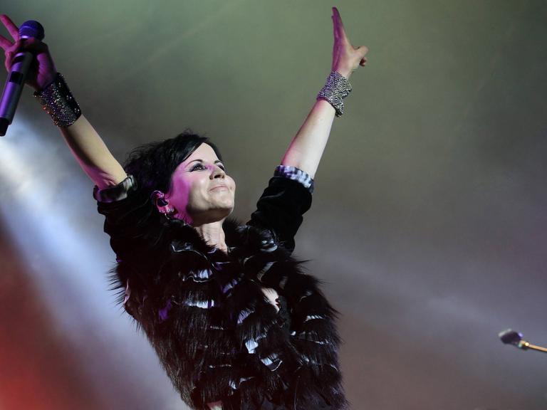 Dolores O’Riordan steht mit nach oben gereckten Armen auf der Bühne, in einer Hand hält sie ein Mikrofon.
