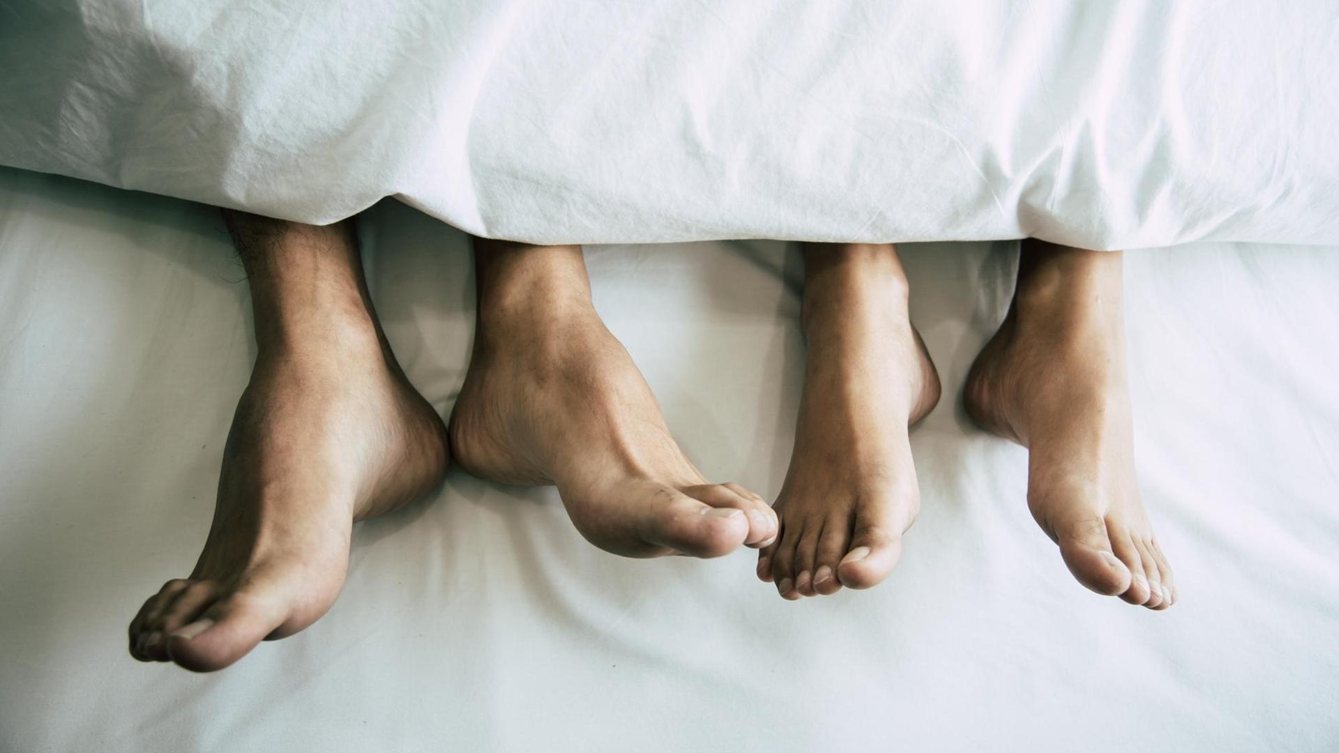 Füße von einer Frau und einem Mann, die unter einer Bettdecke herausschauen.