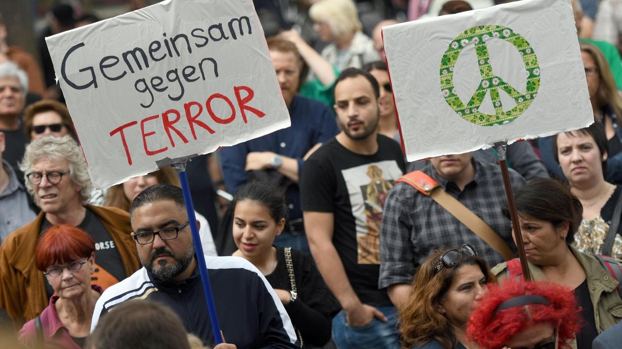 In der Menschenmenge halten ein Mann und eine Frau Schilder mit der Aufschrift "Gemeinsam gegen den Terror" beziehungsweise dem Peace-Symbol hoch.