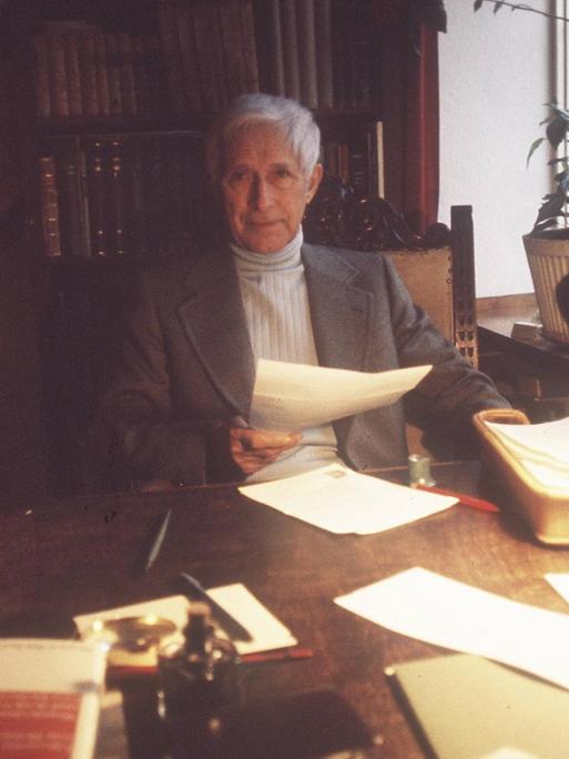 Ein Mann mit kurzen grauen Haaren sitzt an einem Schreibtisch voller Papier