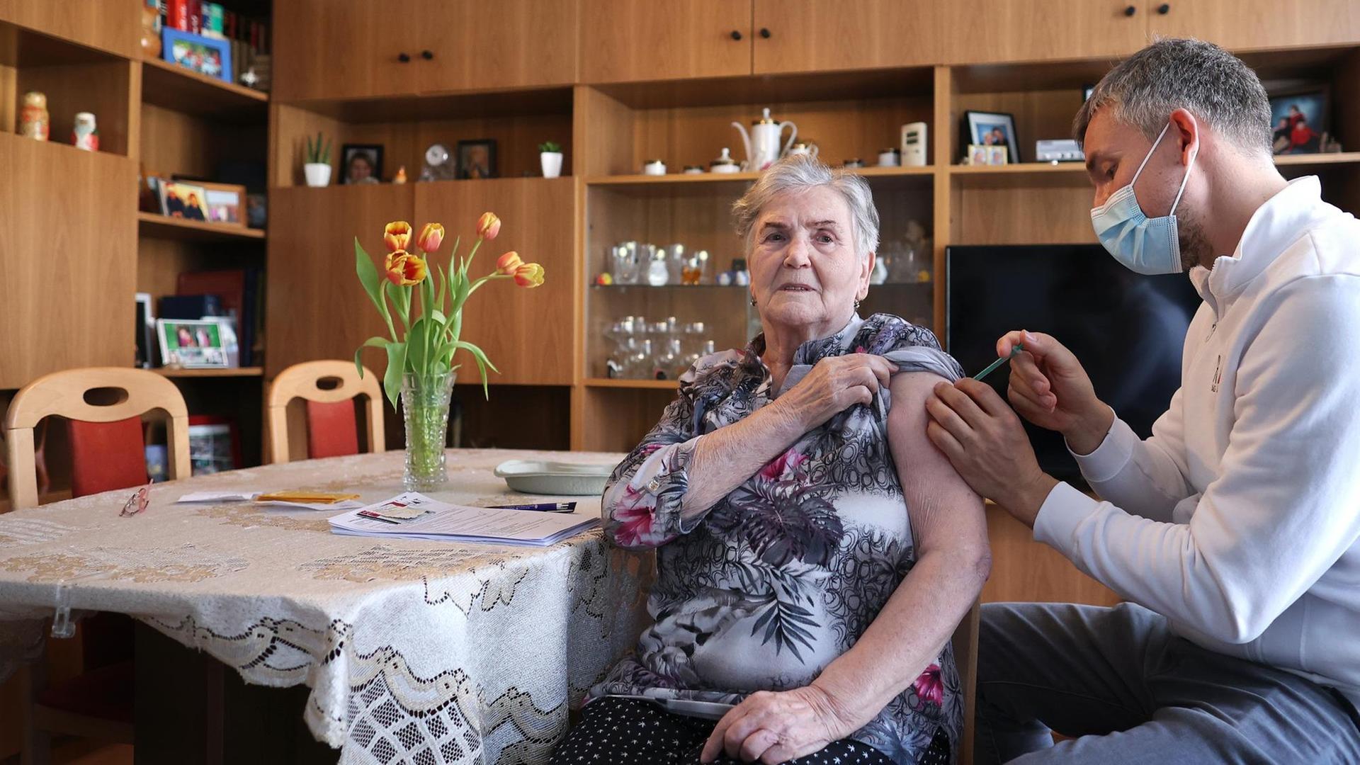 Ein Hausarzt verabreicht einer Seniorin in ihrem Wohnzimmer eine Spritze in den Arm. Ein Hausarzt verabreicht einer Seniorin in ihrem Wohnzimmer eine Spritze in den Arm.