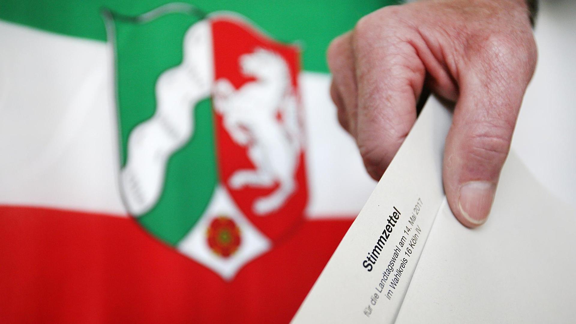 Ein Stimmzettel wird vor der NRW-Fahne in eine Wahlurne gesteckt.