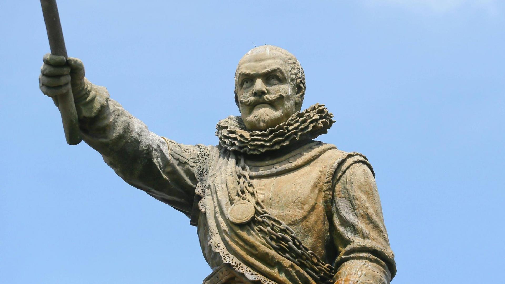 Ein Farbfoto zeigt die Statue eines frühneuzeitlich gekleideten Mannes, der einen starb in den blauen Himmel reckt