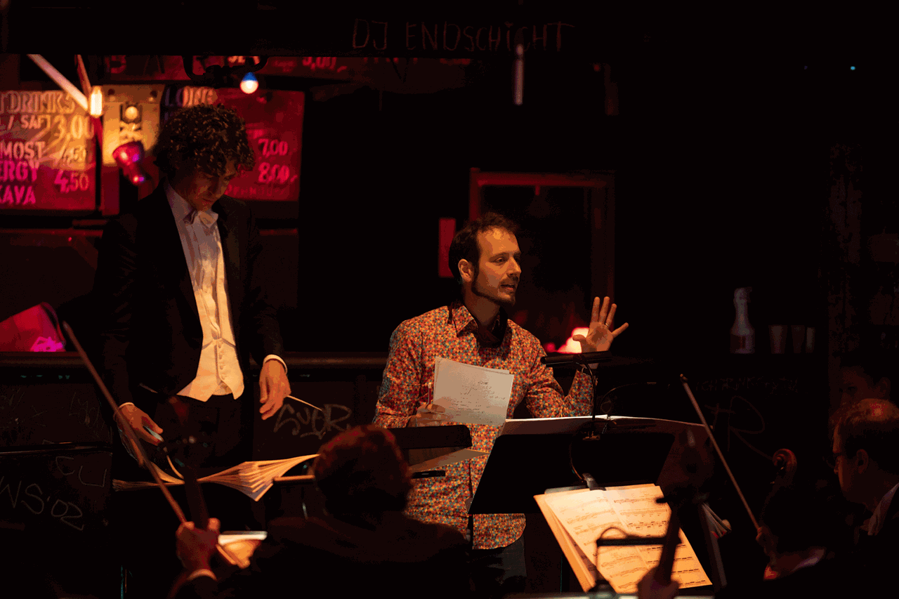 Der Komponist steht mit buntem Hemd in dunkler Clubatmosphäre neben dem Dirigenten des Orchesters und erklärt seine Intensionen.