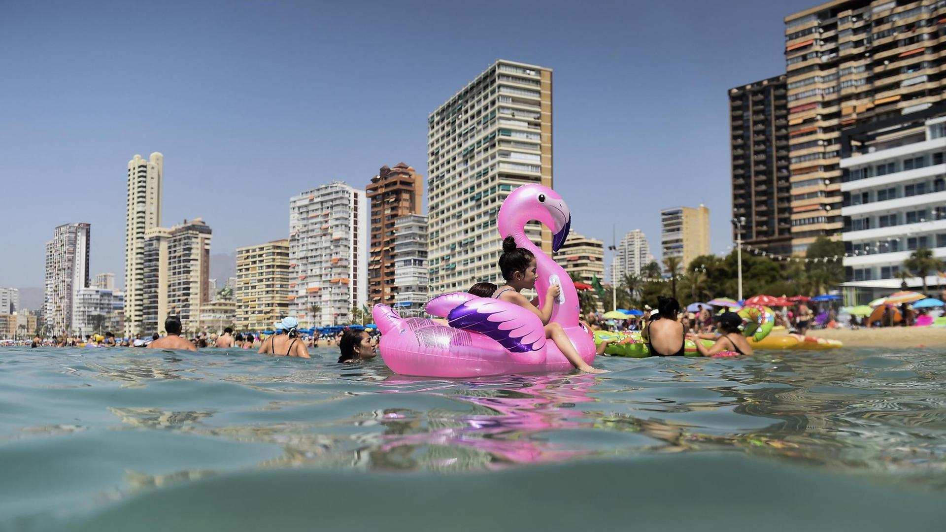 Kinder mit einem rosa Schwimmtier baden im Meer vor den Hochhäusern des Touristenorts Benidorm
