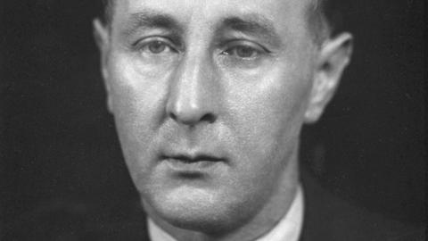 Ein Mann blickt auf einer Schwarzweißfotografie mit etwas ausdrucklosem Blick knapp an der Kamera vorbei.