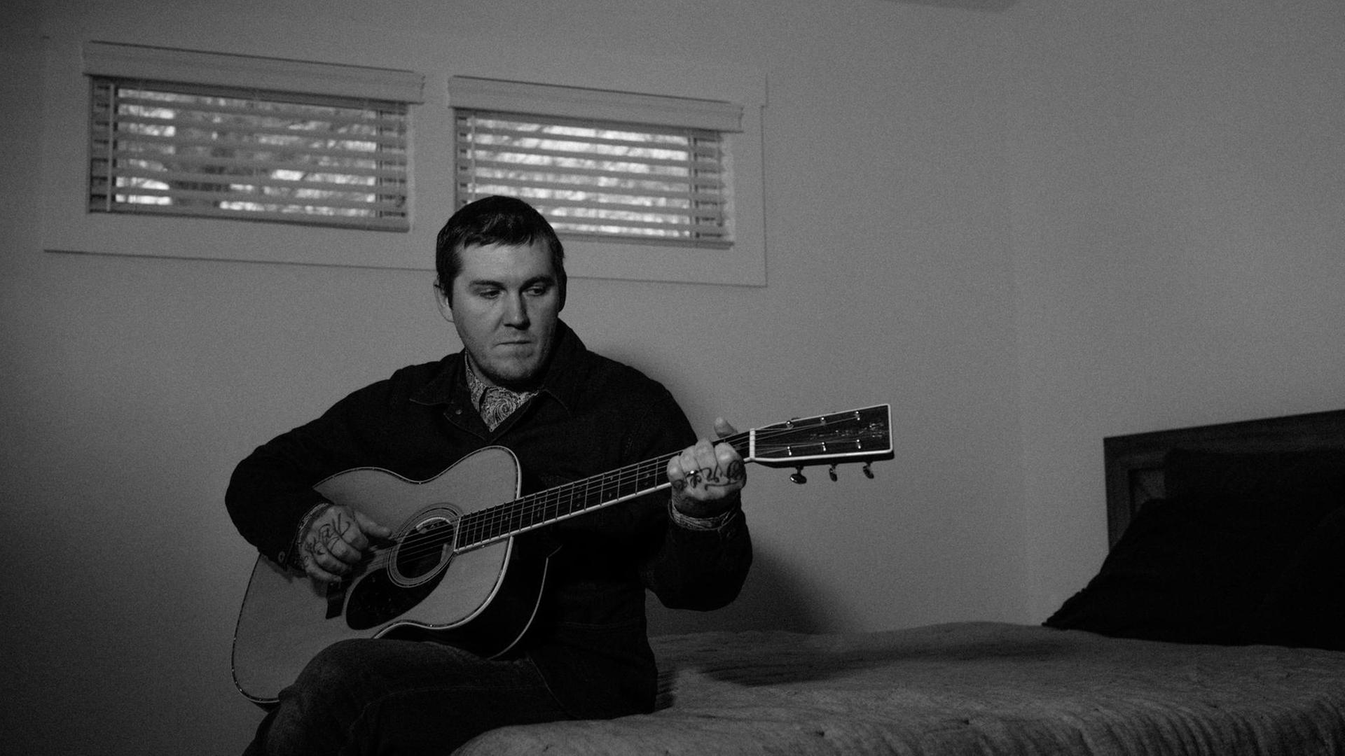 Ein Mann mit kurzen schwarzen Haaren sitzt in einem dunklen Zimmer auf einem Bett und spielt akustische Gitarre.