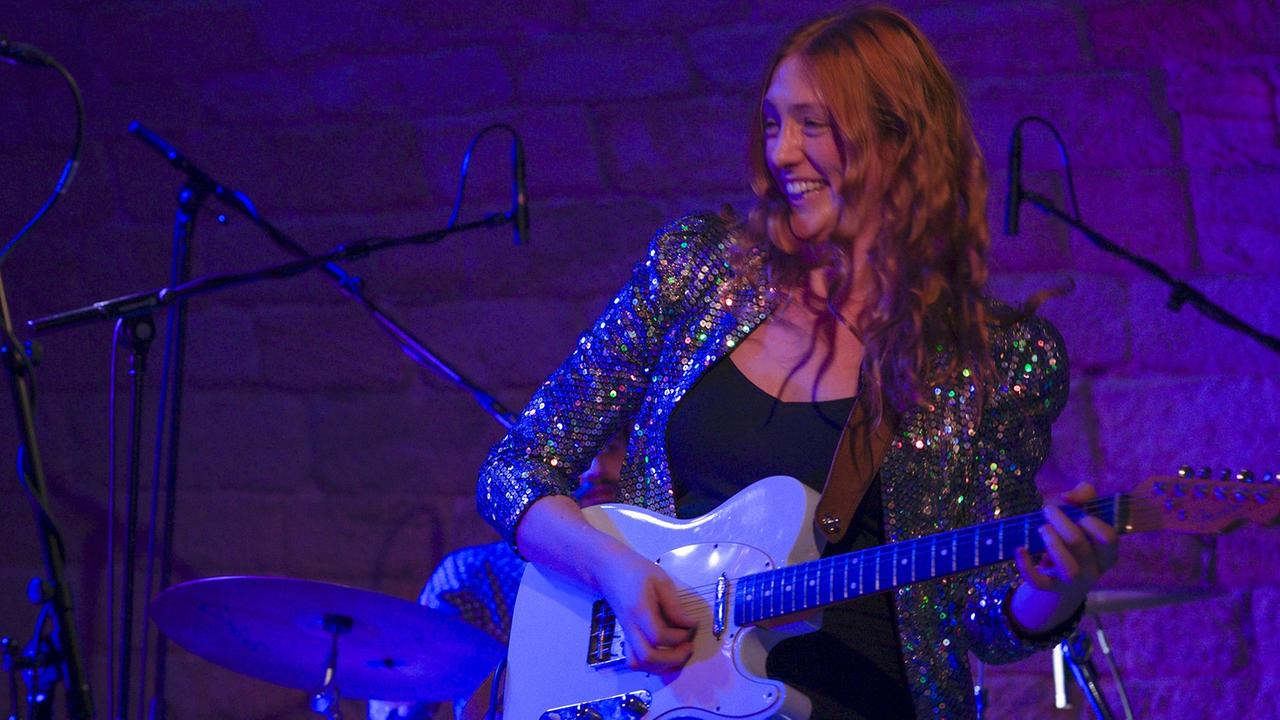 Ein Frau mit langen roten Locken und pailettenbesetztem Jackett steht lächelnd auf einer Bühne und spielt eine weiße E-Gitarre.