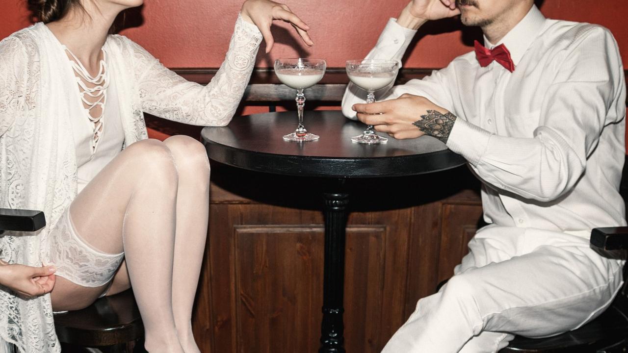Eine Szene in einer Bar. eine weiß gegkleidete Frau und ein ebenfalls weiß gekleideter Mann siezten an einem Tisch und halten Gläster mit weißen Getränken in den Händen. Die Gesichter sind nicht zu sehen.
