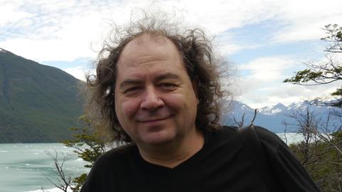 Ein lächelnder Mann mit lockigen Haaren steht vor einem gewaltigen Seen- und Bergpanorama.