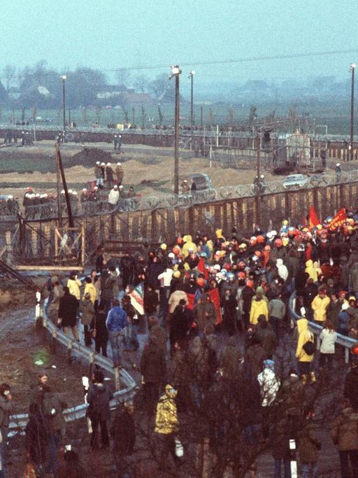 Demonstranten gegen den Kernkraftwerksbau in Brokdorf stehen mit Transparenten vor dem Baugelände, das mit Stacheldrahtzaun und Polizeiaufgebot abgesichert ist. Aufgenommen am 13. November 1976.