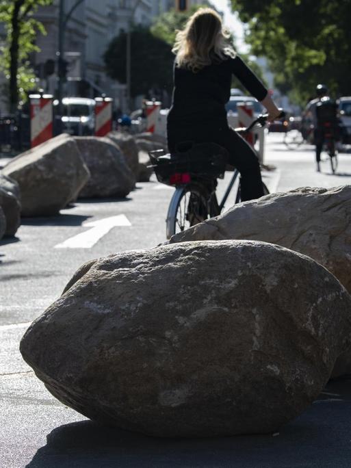 Felsen liegen im Bereich der Kreuzung Bergmannstraße/Zossener Straße auf der Fahrbahn. Eine Fahrradfahrerin schlängelt sich an den Felsbrocken vorbei.