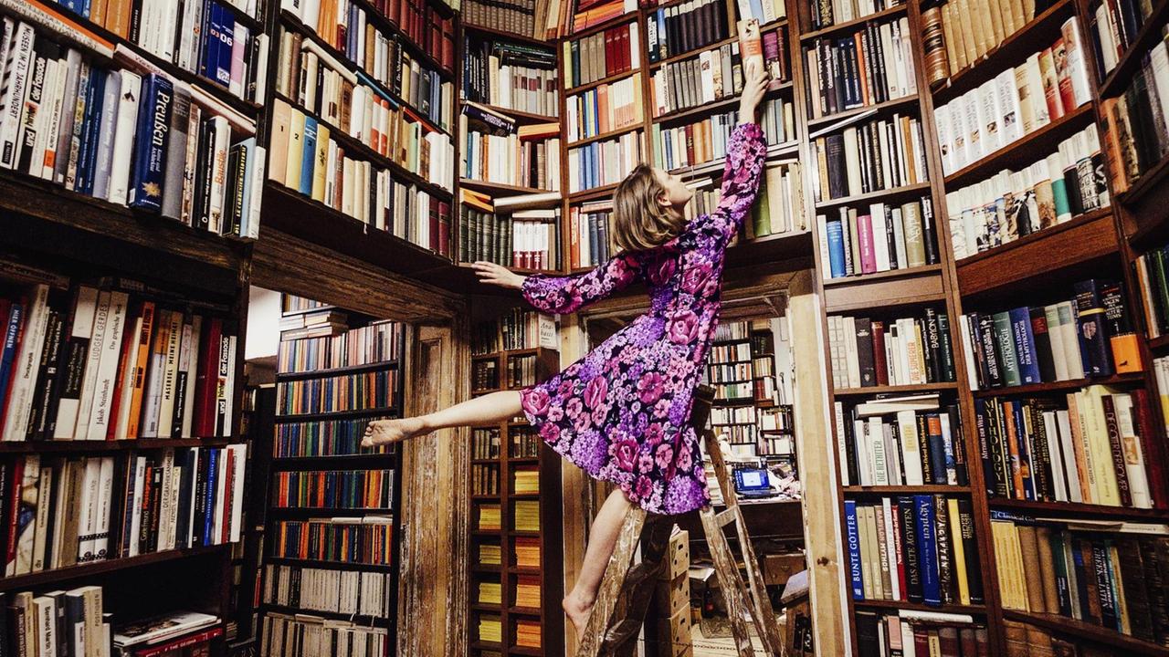 Eine Frau auf einer Leiter angelt ein Buch aus den vollen Regalen einer Bibliothek. Die Bücher nehmen den ganzen Raum ein.