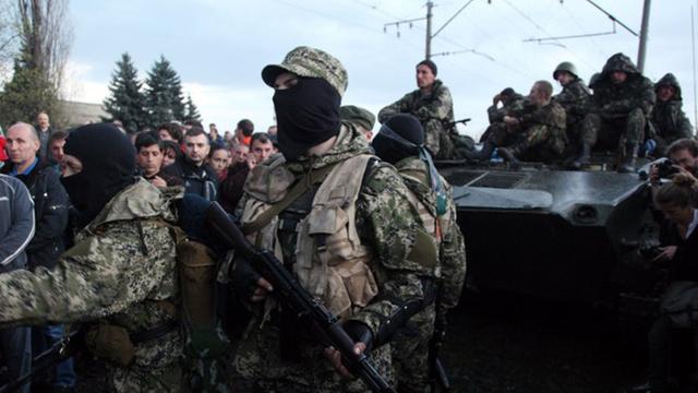 Ein Soldat mit Sturmhaube und Maschinengewehr steht vor einem gepanzerten Fahrzeug, auf dem weitere Soldaten sitzen.
