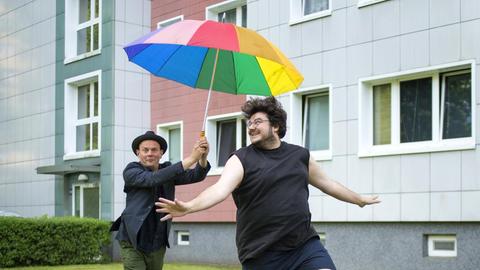 Devid Striesow und Axel Ranisch hüpfend mit einem bunten Regenschirm vor einem Plattenbau.