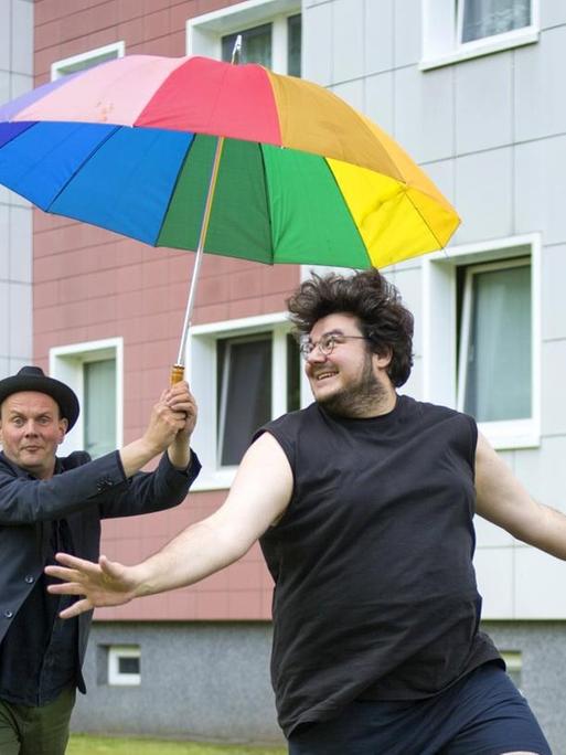 Devid Striesow und Axel Ranisch hüpfend mit einem bunten Regenschirm vor einem Plattenbau.