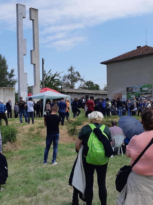 Etwa 100 Menschen versammeln sich auf einer Wiese, dahinter die Ruinen der Gefängnisbaracken