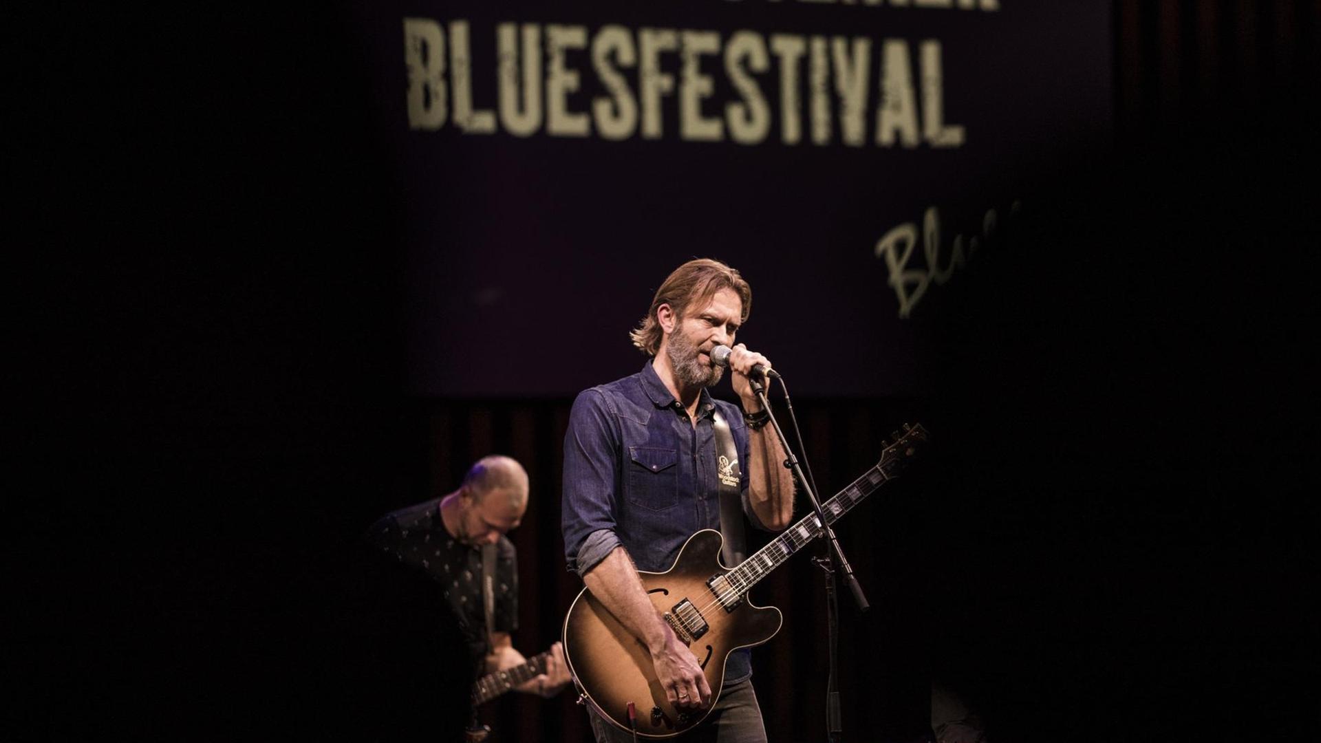 Ein Mann im blauen Hemd steht mit einer Gitarre in der Hand vor einem Mikrofon. Dahinter steht ein weiterer Musiker auf der Bühne.
